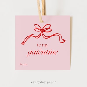 May include: A pink gift tag with a red ribbon design and the words "to my galentine" in red script. The tag also includes the word "from:" and the text "everyday paper". A tan string is attached to the top.