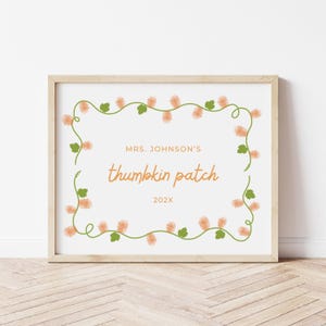 May include: A framed sign with the text "MRS. JOHNSON'S thumbkin patch 202X" in orange script. The sign is bordered by a garland of orange flowers and green leaves on a white background, set in a light wood frame.