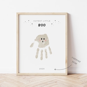 Puede incluir: Una impresi&oacute;n enmarcada con un marco de madera clara. La impresi&oacute;n presenta un dise&ntilde;o de huella de mano en forma de fantasma, con las palabras "Cutest Little Boo" y el a&ntilde;o 202X. El fantasma tiene mejillas rosadas y peque&ntilde;os ojos negros.