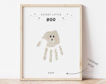 May include: A framed print with a light wood frame. The print features a handprint design in the shape of a ghost, with the words "Cutest Little Boo" and the year 202X. The ghost has pink cheeks and small black eyes.