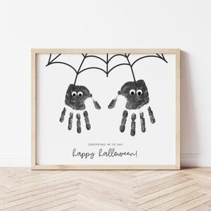 May include: A framed print with two handprint spiders suspended from a black web. The spiders have googly eyes and the text reads "Dropping in to say happy halloween!" The frame is a light wood colour.