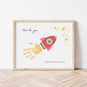 May include: A framed print with a light wood frame. The print features a red rocket with a heart, a handprint, and the words "thank you" and "FOR HELPING ME SOAR" in black text. Yellow stars are scattered across the white background.