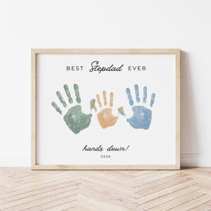 May include: Framed print with the text "BEST Stepdad EVER" above three handprints in green, orange, and blue. The phrase "hands down!" and the year 2026 are below the handprints. The frame is a light wood color.