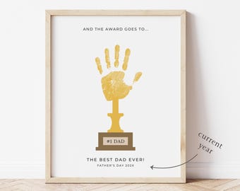 May include: A framed print with a light wood frame. The print features a gold handprint trophy design on a white background. Text includes "AND THE AWARD GOES TO...", "#1 DAD", and "THE BEST DAD EVER!" with "FATHER'S DAY 202X" below. The words "everyday paper" are at the bottom.