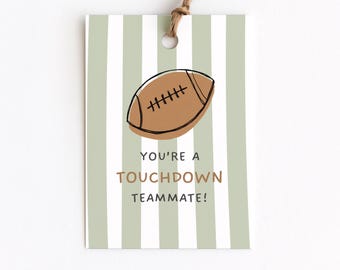 May include: A rectangular paper gift tag with a light sage green and white vertical striped pattern. The tag features a brown illustrated football and the text "YOU'RE A TOUCHDOWN TEAMMATE!" in black and brown lettering, with a brown twine loop.