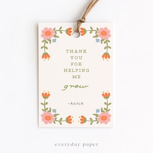 May include: A cream-colored gift tag with a floral border in pink, orange, and blue. The tag reads "THANK YOU FOR HELPING ME grow" with the name "-ANNA" and the text "everyday paper". A brown string is attached.