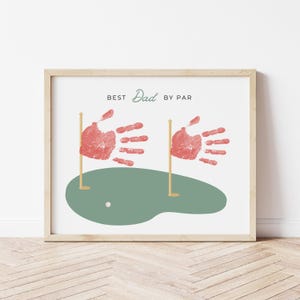 May include: A framed print with the text "BEST Dad BY PAR". The artwork features two red handprints as golf flags, a green golf course, and a white golf ball. The frame is a light wood colour.