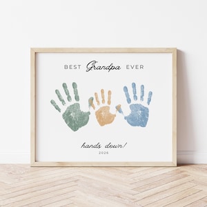 May include: Framed print with the text "BEST Grandpa EVER" and three handprints in green, orange, and blue. The phrase "hands down!" and the year 2026 are also included. The frame is a light wood color.