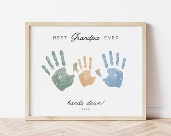 Może przedstawiać: Oprawiony wydruk z napisem "BEST Grandpa EVER" i trzema odciskami dłoni w kolorze zielonym, pomarańczowym i niebieskim. Fraza "hands down!" i rok 2026 są również zawarte. Rama jest w kolorze jasnego drewna.