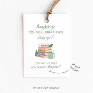 Może przedstawiać: Biała etykieta prezentowa z zielonym napisem "happy SCHOOL LIBRARIAN'S day!". Etykieta przedstawia stos książek, okulary i kubek. Obecny jest również tekst "YOU'RE THE BEST in our book!". Dołączony jest brązowy sznurek.