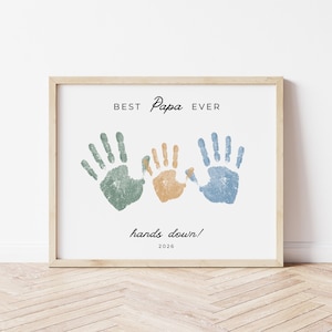 May include: A framed print with the words "BEST Papa EVER" and "hands down!" in black script. Three handprints in green, orange, and blue are below the text. The year 2026 is printed at the bottom.