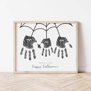 May include: A framed print featuring three handprint spiders hanging from a black web design. The handprints are black with googly eyes. The text "Dropping in to say happy Halloween!" is written in a script font.