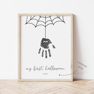 Pode incluir: Impressão emoldurada com um desenho de aranha de impressão manual para o Halloween. A aranha, feita de uma impressão manual com olhos arregalados, está pendurada em uma teia de aranha preta. O texto "my first halloween" e o ano 202X estão abaixo.