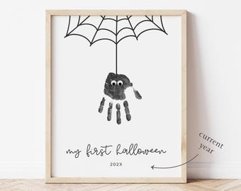 Könnte beinhalten: Gerahmter Druck mit einem Handabdruck-Spinnen-Design für Halloween. Die Spinne, hergestellt aus einem Handabdruck mit Wackelaugen, hängt an einem schwarzen Spinnennetz. Der Text "my first halloween" und das Jahr 202X stehen darunter.