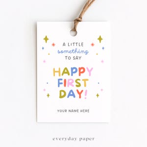 May include: A rectangular white gift tag with a brown twine loop at the top. The text reads A LITTLE something TO SAY HAPPY FIRST DAY! YOUR NAME HERE. The design features colorful star and sparkle icons in yellow, blue, red, and pink.