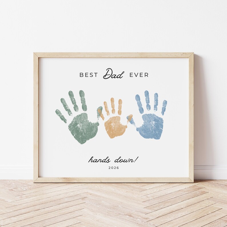 May include: Framed print with the text "BEST Dad EVER" above three handprints in green, orange, and blue. Below the handprints, the text reads "hands down! 2026". The frame is a light wood color.