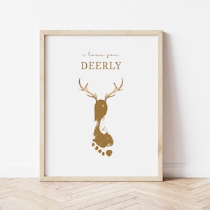 May include: A framed print with a light wood frame. The print features a gold-colored footprint with deer antlers and the words "I love you DEERLY" in a script font. The background is white.