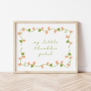 May include: A framed print with the text "my little thumbkin patch" in a whimsical font. The text is surrounded by a border of green vines with peach-colored flower clusters. The frame is a light wood color.