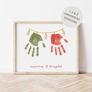 May include: A framed print with a light wood frame. The print features green and red handprints with a string of lights above them. The words "merry & bright" are written in red script below the handprints. The print is on a white background.
