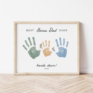 May include: Framed print with the text "BEST Bonus Dad EVER" above three handprints in green, orange, and blue. The phrase "hands down!" and the year 2026 are below the handprints. The frame is a light wood color.