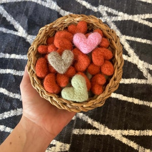 May include: A wicker basket filled with felt hearts in shades of red, pink, and green. The hearts are arranged in a random pattern and are a popular craft project.