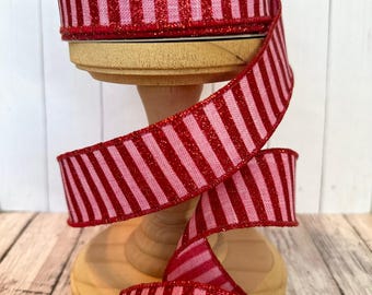 May include: A spool of red and pink striped ribbon with a glittery finish. The ribbon is displayed on a wooden spool holder. The ribbon is a decorative item, suitable for crafts, gift wrapping, or other creative projects.