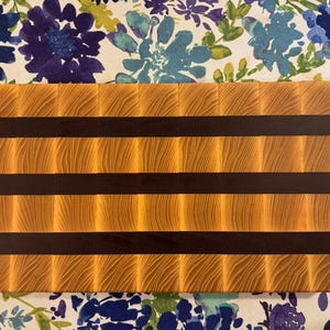 May include: A rectangular end-grain chopping board featuring a repeating pattern of light tan wood blocks with visible grain and two horizontal stripes of dark brown wood. The board has a smooth, flat surface and a clean, geometric construction.