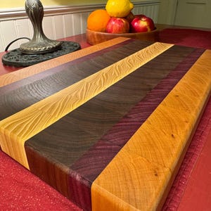 May include: A rectangular end-grain cutting board constructed from alternating strips of wood in varying natural tones, including light tan, dark brown, and deep purple. The surface displays a visible wood grain pattern across the joined segments.