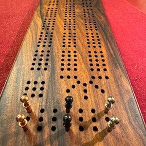 May include: A rectangular wooden cribbage board featuring a dark brown wood grain finish with three parallel tracks of drilled holes. Five metal pegs in gold, black, and silver tones are inserted into the board at the bottom end.