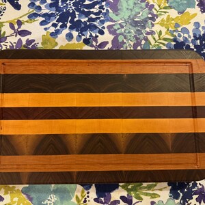 May include: A rectangular wooden chopping board featuring a striped pattern of alternating dark brown, medium brown, and light tan wood segments. The board has rounded corners and a recessed juice groove running along the perimeter of the surface.