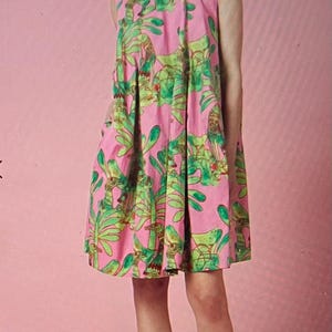 May include: A pink sleeveless dress with a green and pink floral print. The dress has a loose fit and a round neckline. The model is wearing the dress with pink sandals.