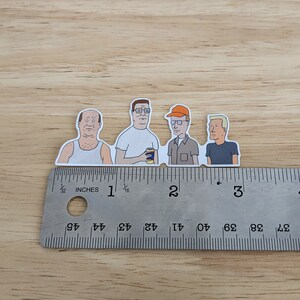 Op de afbeelding: Vier cartoonfiguren uit de animatieserie King of the Hill. De personages staan in een rij op een liniaal met inchmarkeringen. De personages zijn Hank Hill, Dale Gribble, Boomhauer en Bill Dauterive.