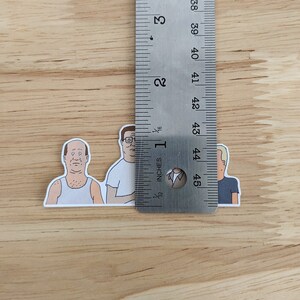 Op de afbeelding: Drie cartoon-personage stickers van mannen uit de animatieserie "King of the Hill". De stickers zijn ongeveer 2,5 cm hoog en zijn gedrukt op wit papier.