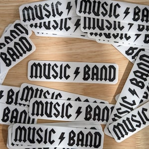 Puede incluir: Una pila de pegatinas blancas con texto negro que dice "Music Band" con un símbolo de rayo entre las palabras. Las pegatinas están dispuestas en círculo sobre una superficie de madera.