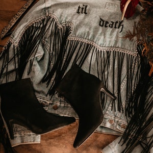 Puede incluir: Una chaqueta vaquera con flecos negros y un detalle de cadena. La chaqueta tiene las palabras "til death" bordadas en la espalda. Dos botas negras de tacón están encima de la chaqueta.