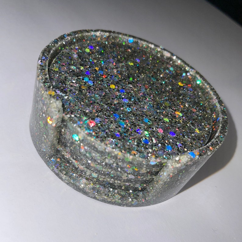 Op de afbeelding: Een set van vier ronde onderzetters met een houder. De onderzetters en de houder zijn gemaakt van een glinsterend, iriserend materiaal met een donkergrijze basis. De glitter reflecteert meerdere kleuren, waaronder blauw, groen en roze.