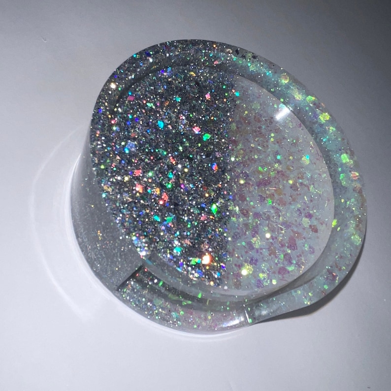 Op de afbeelding: Een ronde asbak van heldere hars met een glinsterend ontwerp. De asbak is gevuld met zilveren en veelkleurige glitter, wat een sprankelend effect cre&euml;ert. Het ontwerp is verdeeld in twee secties, met lichtere glitter aan &eacute;&eacute;n kant.