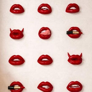 May include: A collection of twelve individual red lip sculptures arranged in a grid. The set features various expressions including lips with small devil horns, a tongue sticking out, lips biting a lipstick tube, and lips biting the lower lip.