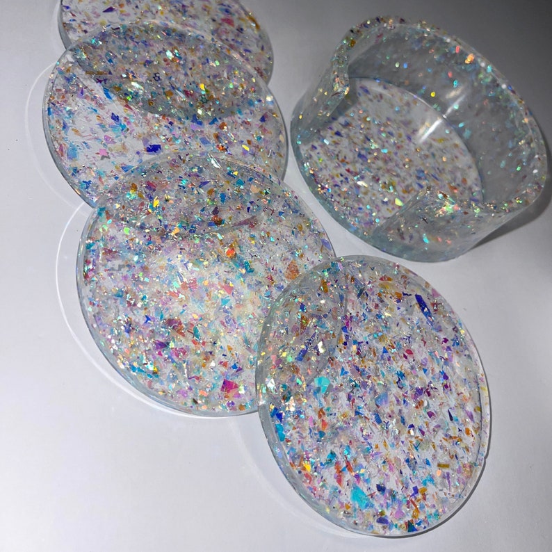 Op de afbeelding: Een set van heldere, ronde onderzetters en een kleine asbak. De onderzetters en asbak zijn gevuld met iriserende, kleurrijke glitterstukjes. De glitter vangt het licht op en cre&euml;ert een sprankelend effect. De onderzetters hebben een diameter van ongeveer 10 cm.