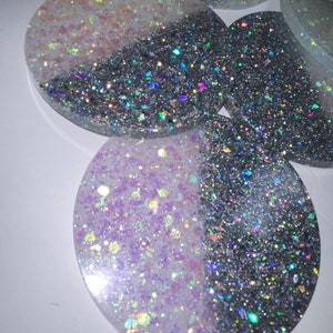 Op de afbeelding: Verschillende ovale onderzetters met een tweekleurig ontwerp. De ene helft is zilverkleurig en glanzend, de andere helft is een mix van witte en iriserende glitter. De onderzetters zijn gemaakt van hars en liggen op een witte ondergrond.