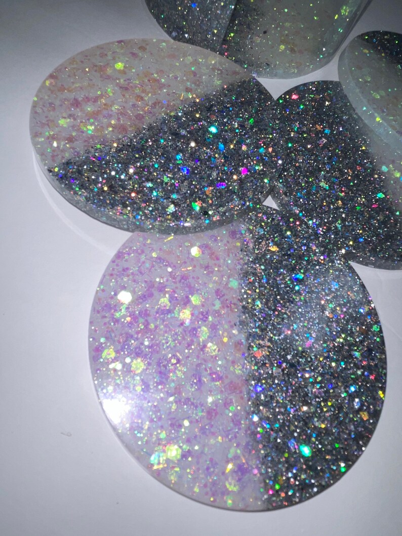 Op de afbeelding: Verschillende ovale onderzetters met een tweekleurig ontwerp. De ene helft is zilverkleurig en glanzend, de andere helft is een mix van witte en iriserende glitter. De onderzetters zijn gemaakt van hars en liggen op een witte ondergrond.