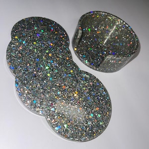 Op de afbeelding: Een set van vier glitterende zilveren hars onderzetters en een bijpassende houder. De onderzetters zijn rond met een unieke vorm, en de houder is een kleine, gebogen schaal. De glitter reflecteert meerdere kleuren.