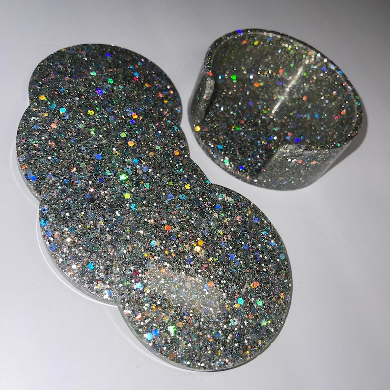 Op de afbeelding: Een set van vier glitterende zilveren hars onderzetters en een bijpassende houder. De onderzetters zijn rond met een unieke vorm, en de houder is een kleine, gebogen schaal. De glitter reflecteert meerdere kleuren.