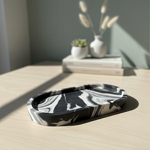 May include: An oval decorative tray with a black and white marble pattern. The tray is made of a solid material and sits on a light-colored wooden surface. The background includes a small white vase with dried flowers and a small succulent in a white pot.