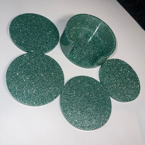 Op de afbeelding: Een set van vijf groene glitter onderzetters en een bijpassende houder. De onderzetters zijn rond en hebben een sprankelende afwerking. De houder is een gebogen, open container. De set is ontworpen om oppervlakken te beschermen tegen drankringen.