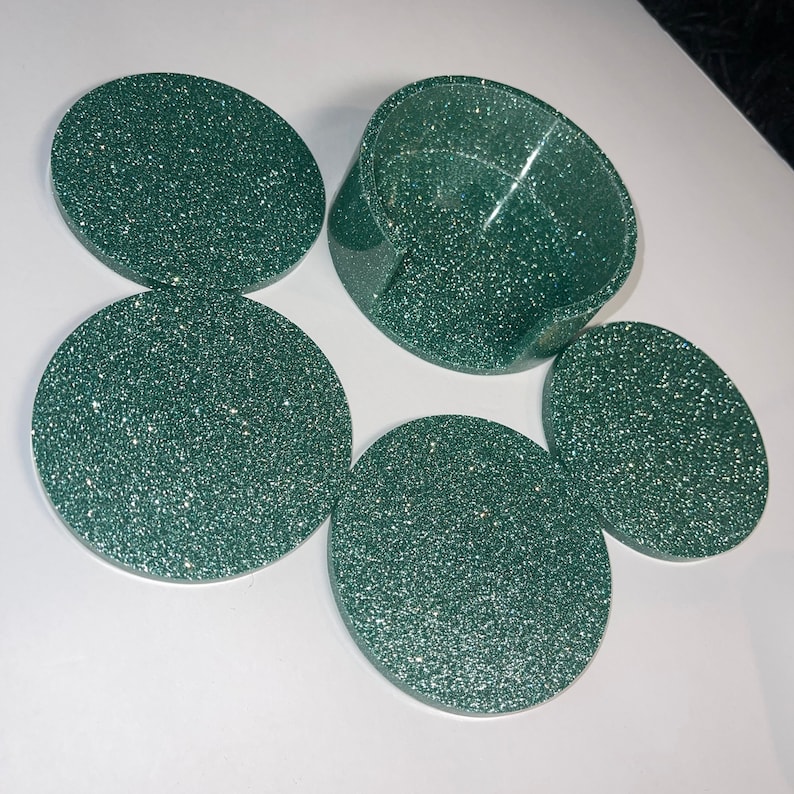 Op de afbeelding: Een set van vijf groene glitter onderzetters en een bijpassende houder. De onderzetters zijn rond en hebben een sprankelende afwerking. De houder is een gebogen, open container. De set is ontworpen om oppervlakken te beschermen tegen drankringen.