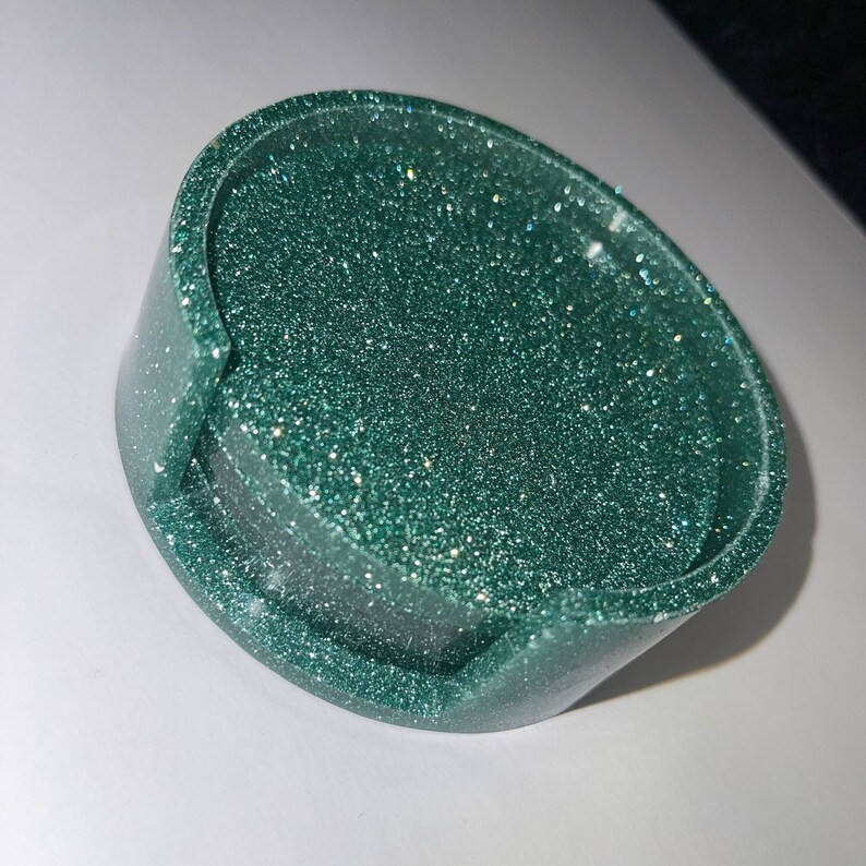 Op de afbeelding: Een ronde, donkergroene onderzetter met ingebedde glitter. De onderzetter heeft een opstaande rand en een verzonken midden. De glitter geeft de onderzetter een sprankelend uiterlijk. Ontworpen om een drankje op te zetten.