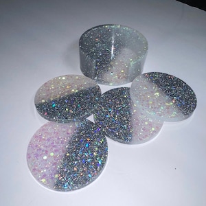 Op de afbeelding: Een set hars onderzetters en een bijpassende houder. De onderzetters zijn rond, met een gesplitst ontwerp van glinsterend zilver en wit of roze. De houder is een korte cilinder, eveneens met een zilver en wit glitterontwerp.