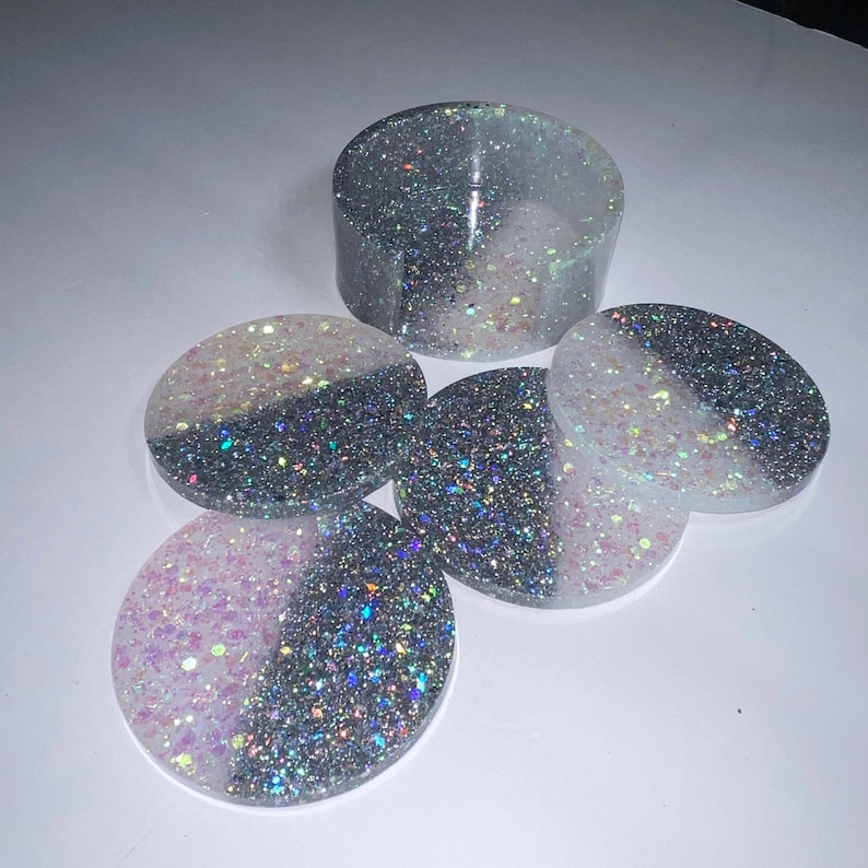 Op de afbeelding: Een set hars onderzetters en een bijpassende houder. De onderzetters zijn rond, met een gesplitst ontwerp van glinsterend zilver en wit of roze. De houder is een korte cilinder, eveneens met een zilver en wit glitterontwerp.