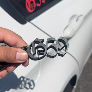 May include: A dark gray 3D-printed keychain featuring the number "659" in a gothic font. The keychain is held in front of a white car door. The number "659" is also visible in red on the car.